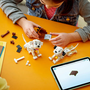 Child assembling LEGO 101 Dalmatians Lucky and Penny puppy figures on bright yellow surface with scattered LEGO bricks, instruction manual and tablet displaying building guide