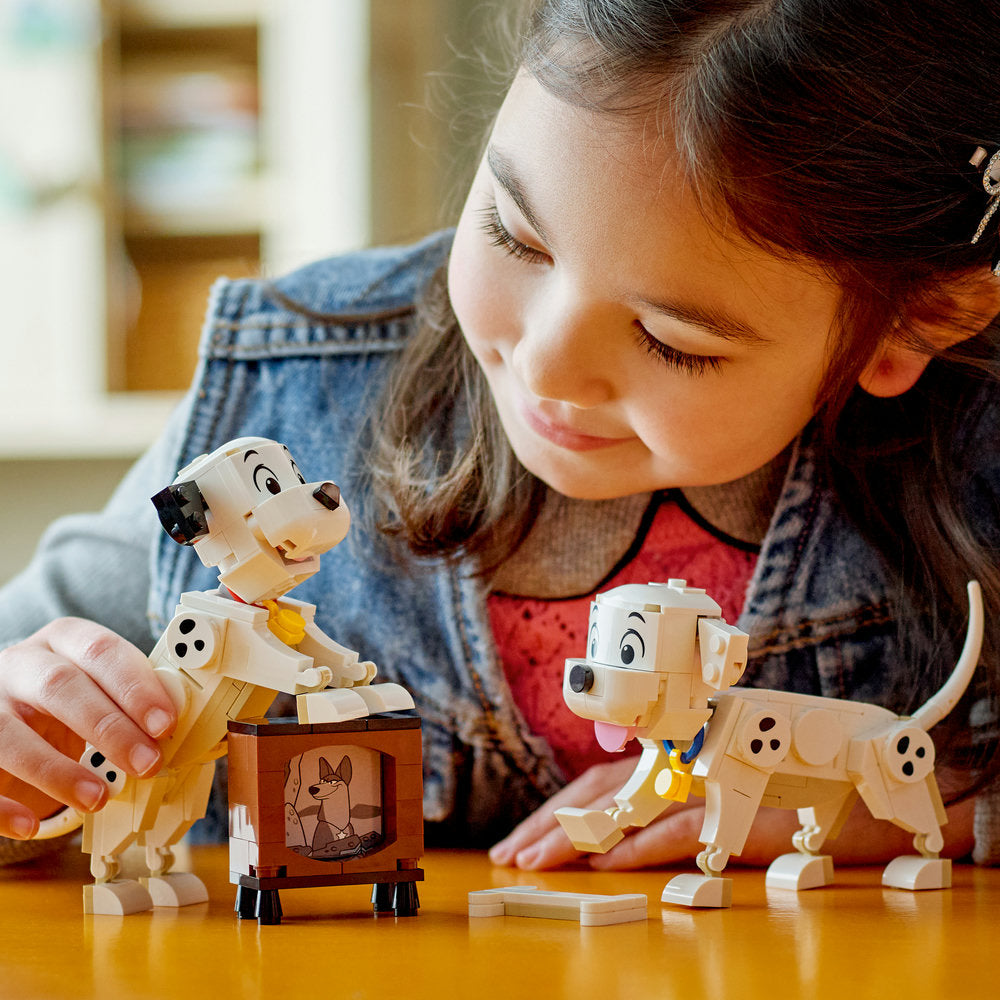 Young girl playing with LEGO Disney 101 Dalmatians Lucky and Penny puppy figurines on wooden table, featuring tan and white buildable dog characters with spots and a brown kennel accessory piece