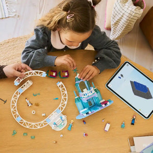 Young child playing with LEGO Elsa's Ice Castle set 43281 at wooden table, featuring blue ice castle structure, white roller coaster track, minifigures, and instruction display on tablet