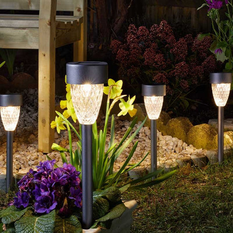 Four solar garden stake lights with textured glass shades and dark metal stems illuminating a garden bed at night, surrounded by purple petunias, yellow flowers, burgundy seed heads, ornamental grasses, and decorative gravel near wooden decking