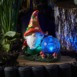 Colorful garden gnome solar light with red pointed hat decorated with yellow flowers, white beard, blue glowing crystal ball, green outfit and mushroom base, displayed in a garden setting against weathered stone wall