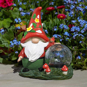 Garden gnome solar light with red pointed hat decorated with yellow flowers and white mushrooms, white beard, green outfit, holding a dark mosaic gazing ball surrounded by small red and white mushrooms, displayed among blue forget-me-nots and red flowers in a garden setting