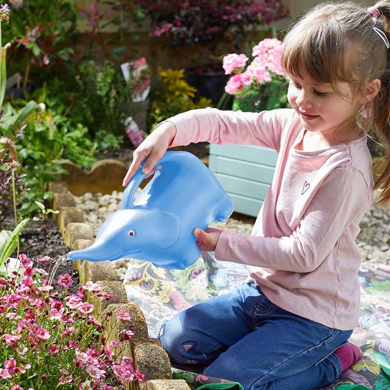 Young girl wearing pink cardigan watering pink flowers in a garden with a blue elephant-shaped watering can