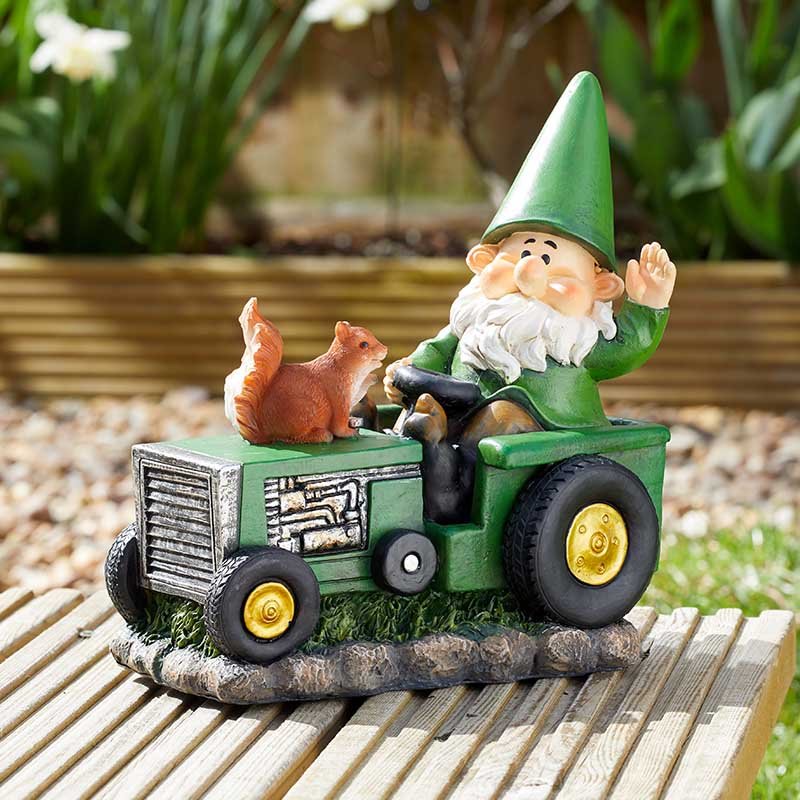 Garden gnome figurine wearing green pointed hat and beard sitting in green tractor with yellow wheels next to brown squirrel, positioned on wooden decking with garden background