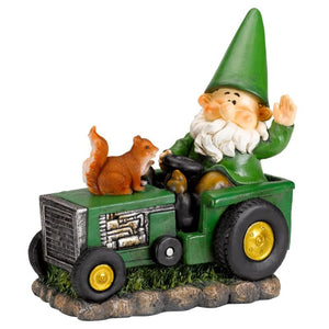 Garden gnome figurine wearing green pointed hat sitting on green tractor with yellow wheels, accompanied by brown squirrel, featuring white beard and waving hand, displayed on rocky base