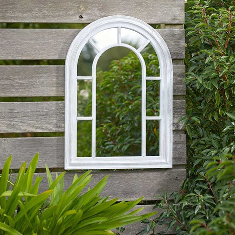 White arched garden mirror mounted on grey wooden fence surrounded by green plants and foliage