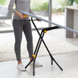 Woman using grey and yellow Joseph Joseph Glide ironing board with black X-frame legs on light wooden floor in modern living room with large windows