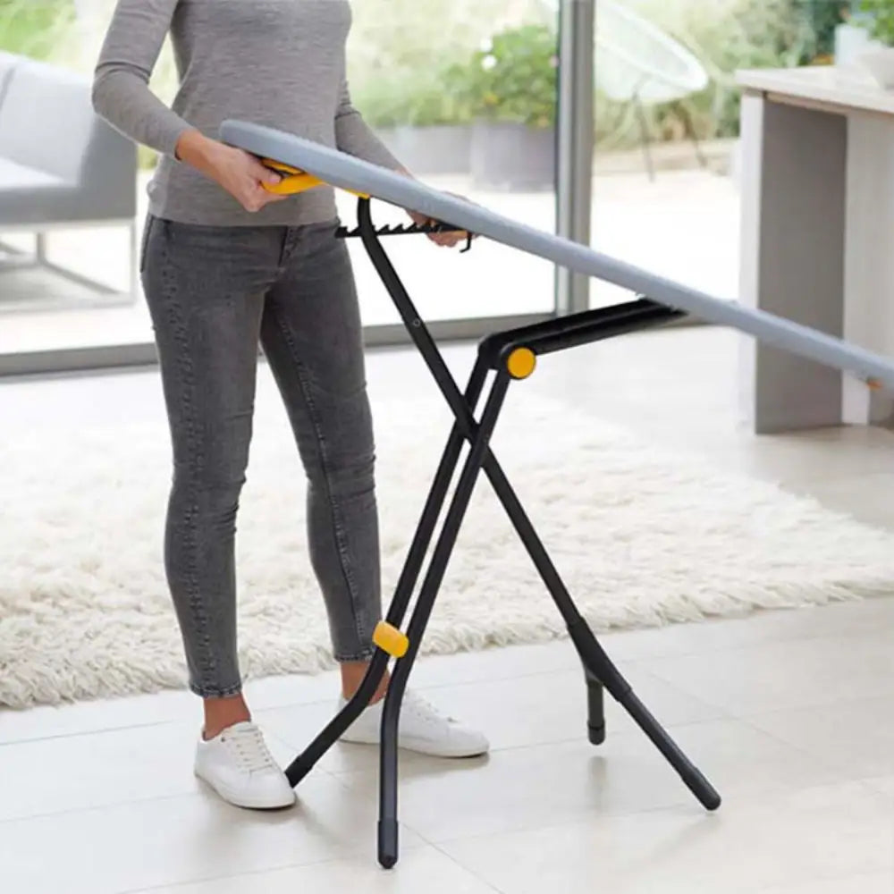 Woman using grey and yellow Joseph Joseph Glide ironing board with black X-frame legs on light wooden floor in modern living room with large windows