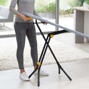 Woman using grey and yellow Joseph Joseph Glide ironing board with black X-frame legs on light wooden floor in modern living room with large windows
