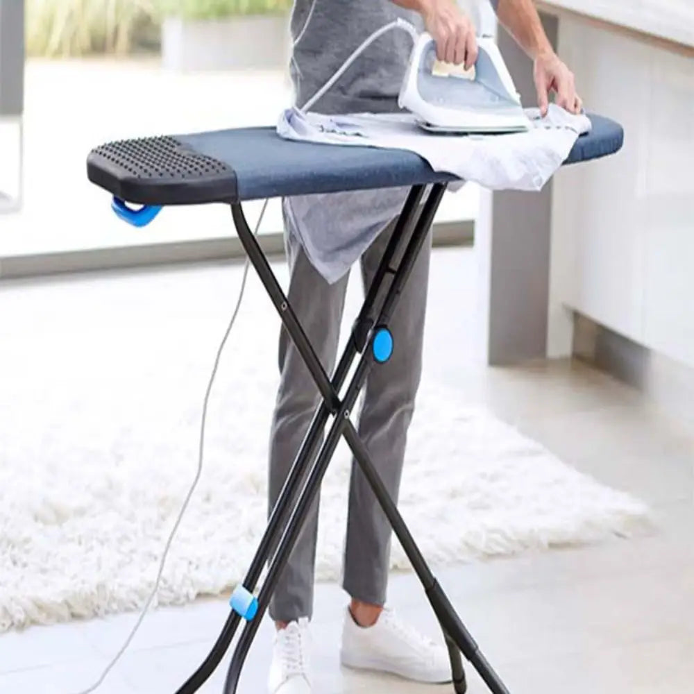 Person using a compact Joseph Joseph Glide ironing board with black metal X-frame legs, blue accents, and grey padded ironing surface, while pressing a white shirt indoors