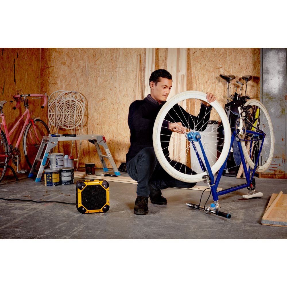 Man working on blue bicycle in workshop holding white wheel, with yellow and black Dimplex Rugged electric fan heater on concrete floor, surrounded by tools and bikes against cork walls