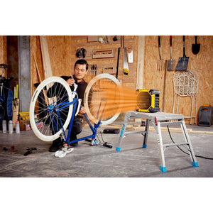 Man in workshop holding bicycle wheel next to blue bike frame on stand with Dimplex yellow and black industrial electric fan heater on white ladder, cork wall background with tools