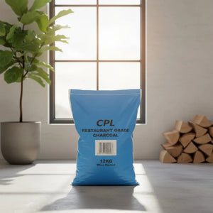 Blue CPL Restaurant Grade Charcoal 12kg bag displayed on a light gray surface near a potted green plant, windows, and stacked firewood in a minimalist interior setting