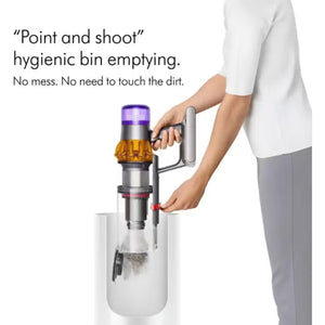 Person demonstrating hygienic bin emptying feature of Dyson V15 Detect vacuum with point and shoot mechanism, showing stainless steel dustbin with purple top cap and red release button, dirt visible in collection chamber below