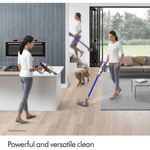 Woman using Dyson V11 cordless vacuum cleaner with purple and blue design across modern kitchen, hallway and living room spaces, demonstrating versatile cleaning on wooden floors and carpets with dog nearby