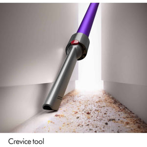 Close-up of Dyson V11 Advanced cordless vacuum cleaner with purple handle and silver-gray motorized head with red indicator light, shown cleaning debris from white floor with crevice tool attachment labeled below