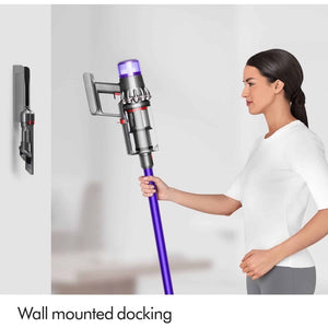Woman in white shirt demonstrating Dyson V11 Advanced cordless vacuum with purple and silver body docked on wall-mounted charging station in minimalist white interior