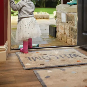 Young child in gray hoodie and pink leggings standing on beige Hug Rug runner mat with footprint pattern at open doorway leading to garden patio