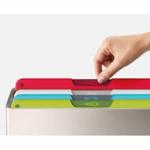 Hand pulling red chopping board from stacked Joseph Joseph Folio Icon multicolored cutting board set showing red, white, blue, gray and green boards with integrated handles and icons