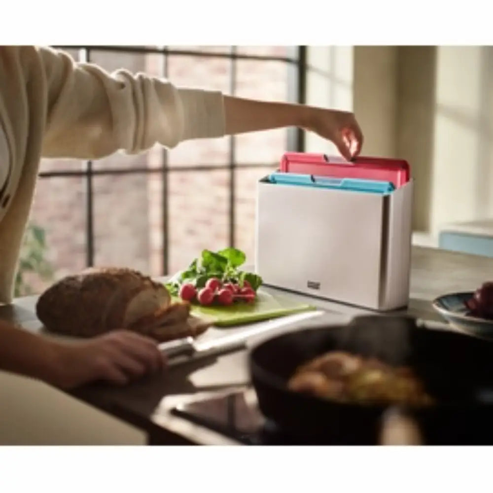 Person using a Joseph Joseph Folio Icon chopping board set with pink, turquoise, and white boards stacked together on a kitchen counter, with bread, radishes, and vegetables visible during food preparation