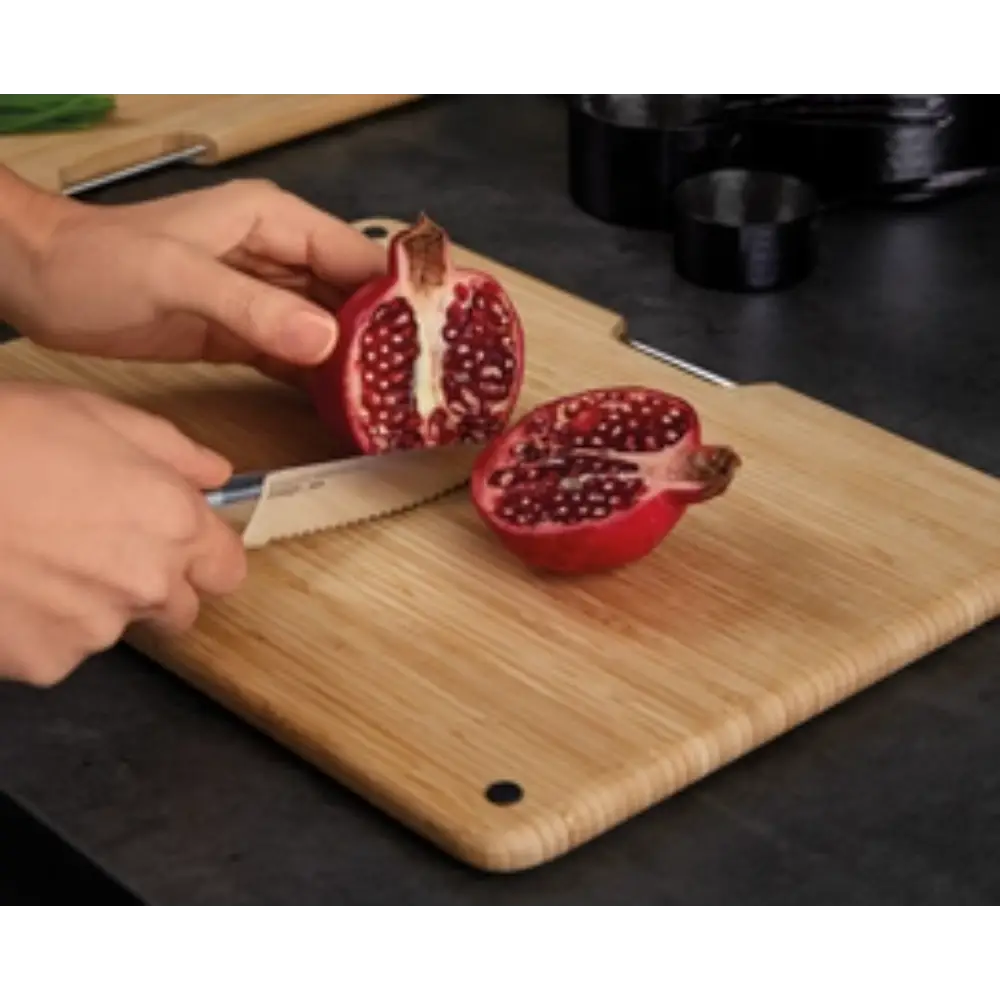 Hands demonstrating cutting a pomegranate in half on a natural bamboo chopping board from the Joseph Joseph Folio set, with a serrated steel knife, showing the red fruit's seeds against the light wood surface in a kitchen setting