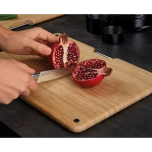 Hands demonstrating cutting a pomegranate in half on a natural bamboo chopping board from the Joseph Joseph Folio set, with a serrated steel knife, showing the red fruit's seeds against the light wood surface in a kitchen setting