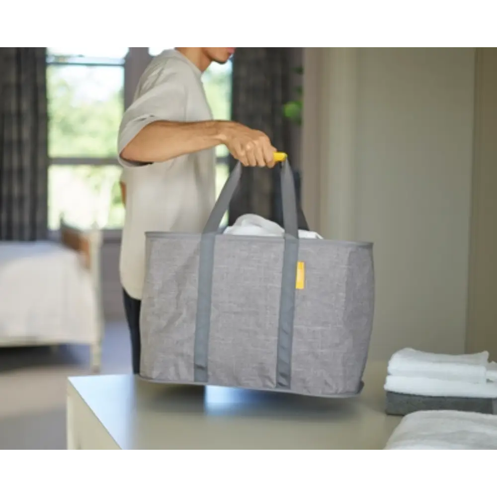 Person holding a grey fabric laundry basket with handles in a modern bedroom setting, demonstrating the Joseph Joseph Hold All Max Laundry Basket with yellow accent details