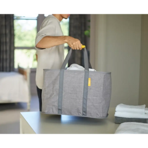 Person holding a grey fabric laundry basket with handles in a modern bedroom setting, demonstrating the Joseph Joseph Hold All Max Laundry Basket with yellow accent details