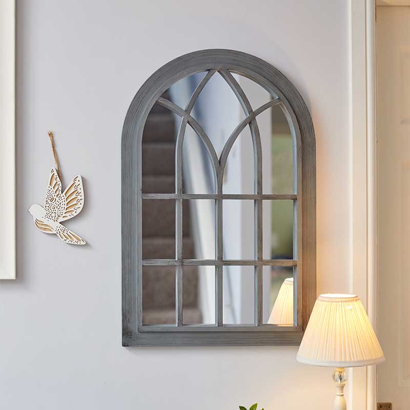 Slate grey arched window-style mirror with geometric pane detailing hanging on white wall beside decorative gold leaf ornament and cream table lamp