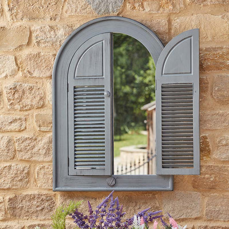 Slate grey arched louvre window mirror mounted on sandstone brick wall with purple and pink flowers below, reflecting green garden and outdoor scenery