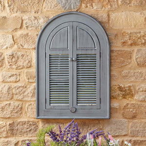 Slate grey arched louvre window mirror mounted on honey-coloured stone wall with purple and white flowering plants below