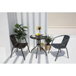 A 2 seater balcony furniture set comprising of two stackable chairs and a round table with a glass top, all in dark grey color, placed on a balcony with plants in the background.