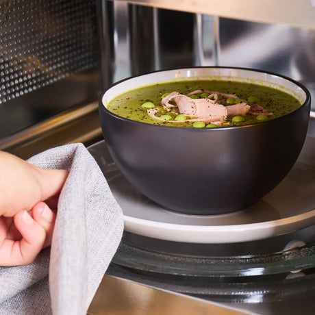 Grey Flint Morphy Richards microwave with black bowl of green soup containing meat and peas being removed with cloth, showing microwave interior and stainless steel details