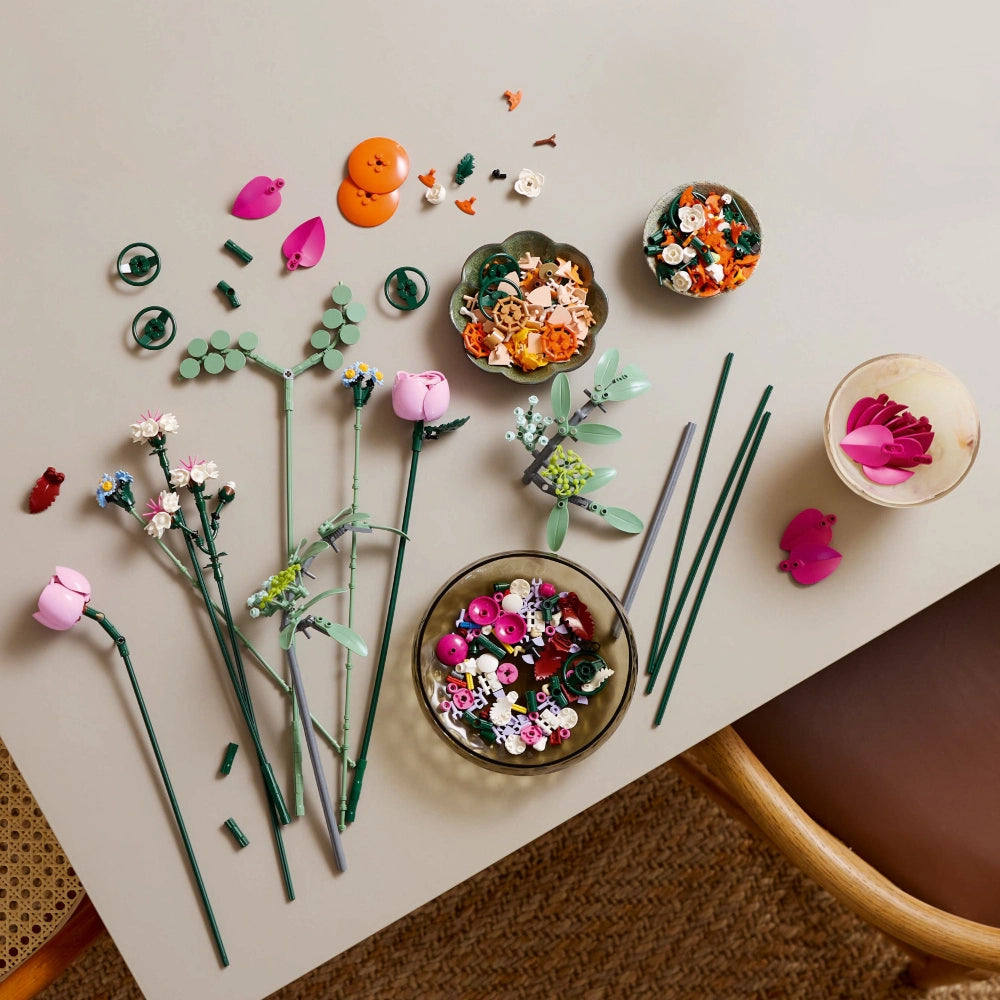 Flat lay overhead view of colorful LEGO flower building set components arranged on beige surface, including pink roses, orange blooms, white flowers, green stems and leaves, decorative bowls with assembled flower arrangements, and small decorative elements in pink, orange, and green tones
