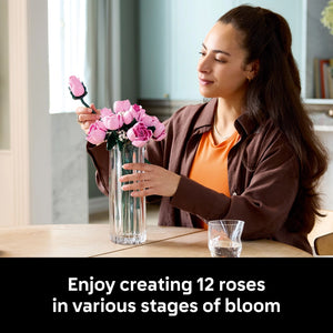 Woman arranging pink LEGO roses in a clear glass vase, wearing a brown jacket over an orange top, displaying 12 buildable roses in various stages of bloom