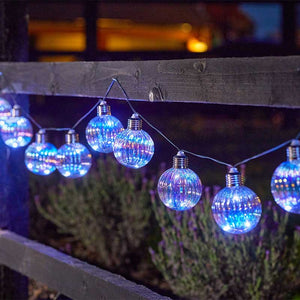 String lights with ten blue LED orb bulbs hanging from wire against wooden fence with lavender plants below and warm building lights blurred in background at dusk