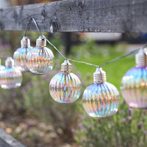 Smart Garden Firefly Opal Orb Garden String Lights with 10 iridescent ribbed glass globe bulbs suspended from a wire cable strung across a garden setting with green grass and stone wall in background