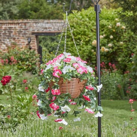Regal hanging basket with cascading pink and white petunia flowers in woven brown basket suspended from chain in lush garden setting with brick wall and flowering plants in background