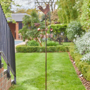 Copper cockerel weather vane with directional arrow and cardinal points mounted on tall pole in garden setting with green lawn and hedges