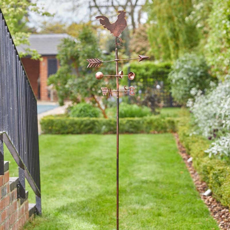 Copper cockerel weather vane with directional arrow and cardinal points mounted on tall pole in garden setting with green lawn and hedges