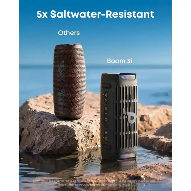 Soundcore Boom 3i black portable Bluetooth speaker positioned on wet sand next to rocks with ocean backdrop, highlighting 5x saltwater-resistant construction compared to competitor products