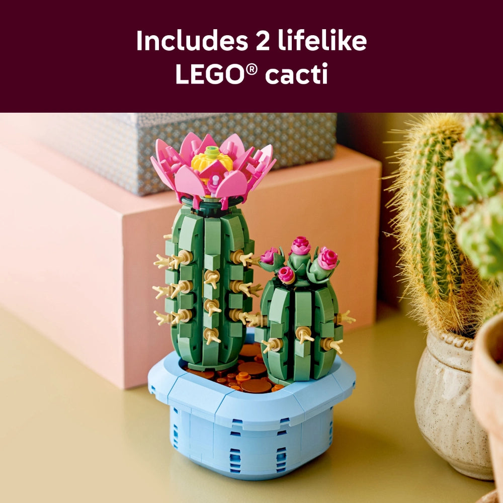 LEGO Flowering Cactus building set featuring two lifelike green cacti with pink and red blooms and tan spines in a light blue pot, displayed on a yellow surface surrounded by real potted plants and neutral-toned decorative boxes