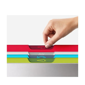 Hand inserting a red chopping board into a Joseph Joseph Folio 4-piece color-coded chopping board set showing stacked boards in red, white, blue, yellow-green and gray with fish icons