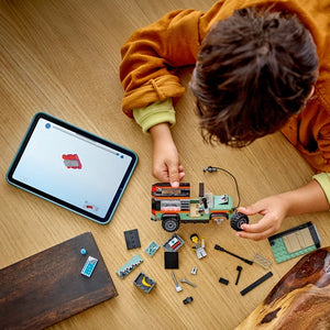 Child in orange hoodie assembling LEGO Off-Road 4x4 Mountain Truck set on wooden floor with tablet displaying instructions, showing orange and black truck model and loose LEGO bricks and pieces