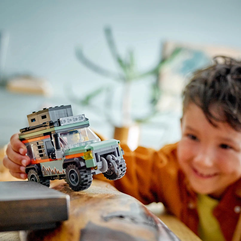 Young child in mustard yellow shirt playing with LEGO Off-Road 4x4 Mountain Truck set featuring green and orange buildable truck with detailed construction blocks stacked on cargo bed, displayed on sandy surface with blurred potted plant in background