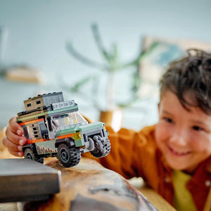 Young child in mustard yellow shirt playing with LEGO Off-Road 4x4 Mountain Truck set featuring green and orange buildable truck with detailed construction blocks stacked on cargo bed, displayed on sandy surface with blurred potted plant in background