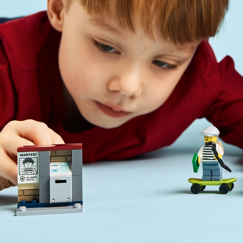 Young child in red shirt playing with LEGO Police Motorcycle Chase set 60455, featuring a wanted poster police station building and minifigure on yellow skateboard against light blue background