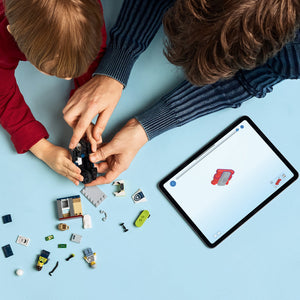 Two children assembling LEGO Police Motorcycle Chase set 60455 on a light blue surface, with hands holding black motorcycle piece above scattered colorful LEGO components and a tablet displaying the red police motorcycle build instructions