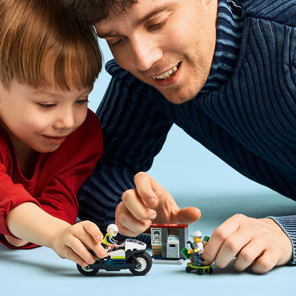An adult and child playing together with a LEGO Police Motorcycle Chase set 60455, featuring a black and yellow motorcycle, police station building, and minifigures against a blue background