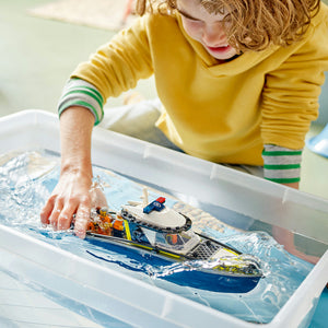 Child in yellow shirt playing with LEGO Police Boat Chase set 60456 in water-filled white bin, featuring blue and white police speedboat with minifigures and orange getaway boat